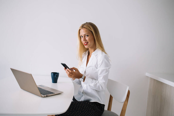 Concentrated young blonde woman sitting at white table with blue cup in blouse texting on smartphone using laptop and looking at camera - Foto, imagen
