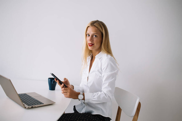 Content young blonde woman sitting at white table with blue cup in blouse texting on smartphone using laptop on white background and looking at camera - Foto, Imagen