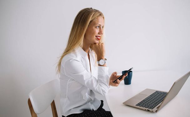 Side view of concentrated smiling young blonde woman sitting at white table with blue cup in blouse texting on smartphone using laptop on white background - Foto, afbeelding