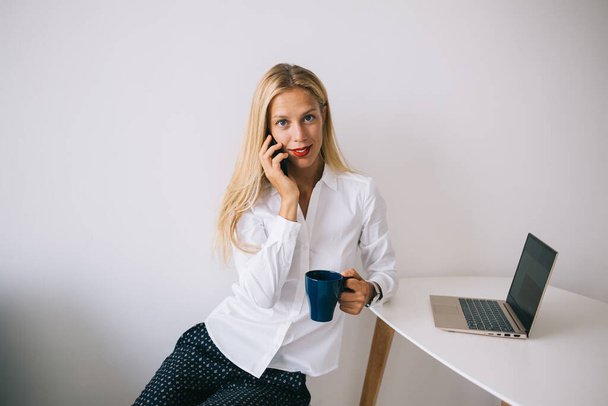 Concentrated happy young blonde woman leaning on white table with blue cup in blouse speaking on smartphone using laptop and looking at camera on white background - Foto, immagini