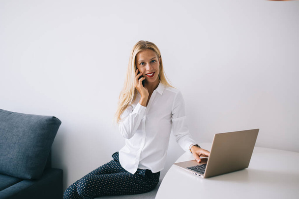Satisfied young blonde woman sitting at white table in blouse speaking on smartphone using laptop at home and looking at camera - Фото, зображення
