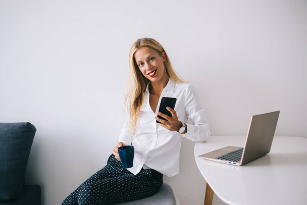 Satisfecho feliz joven mujer rubia inteligente apoyándose en la mesa blanca manteniendo taza azul en blusa mensajes de texto en el teléfono inteligente utilizando el ordenador portátil en casa y mirando a la cámara - Foto, imagen