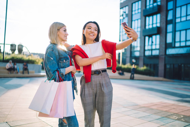 Pleased young multiracial charming female friends with shopping bags taking selfie on smartphone while standing near mall in urban street - Foto, Bild