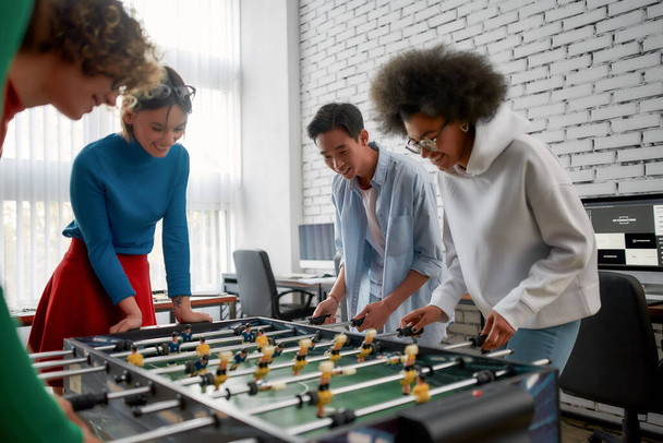 A termelékenység növelése. Fiatal és boldog multikulturális emberek alkalmi viseletben asztali fociznak a modern irodában. Irodai tevékenységek. Jól szórakozunk együtt. Boldog alkalmazottak - Fotó, kép