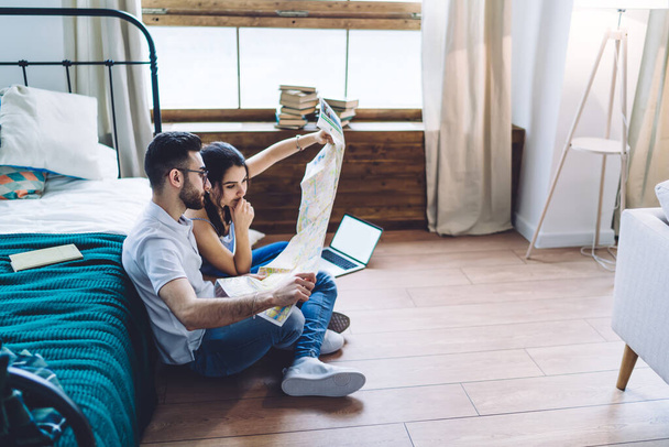 Side view of concentrated casual couple preparing for trip and exploring map together while sitting on floor with laptop beside bed in modern apartment - Fotoğraf, Görsel