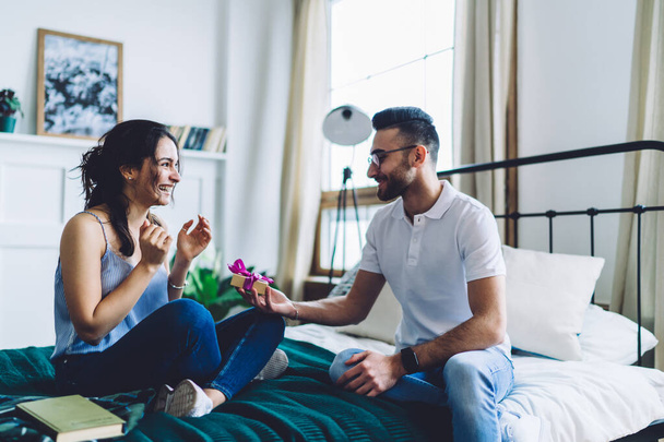 Excited shy smiling boyfriend proposing happy emotional positive woman with ring in beautiful small box sitting together on bed in bedroom  - Foto, afbeelding