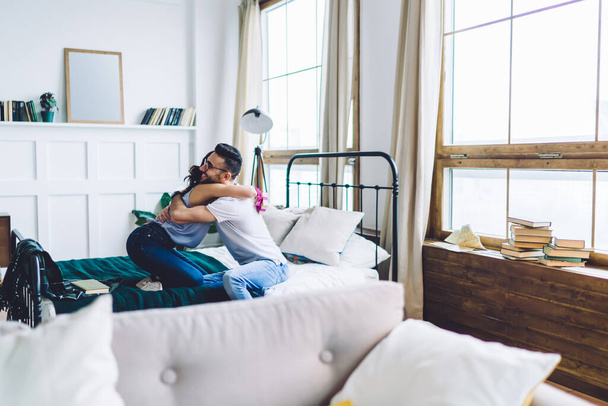 Zijaanzicht van jonge liefdevolle liefdevolle sensuele paar zitten op bed en knuffelen na het geven van cadeau in gezellige moderne appartement - Foto, afbeelding