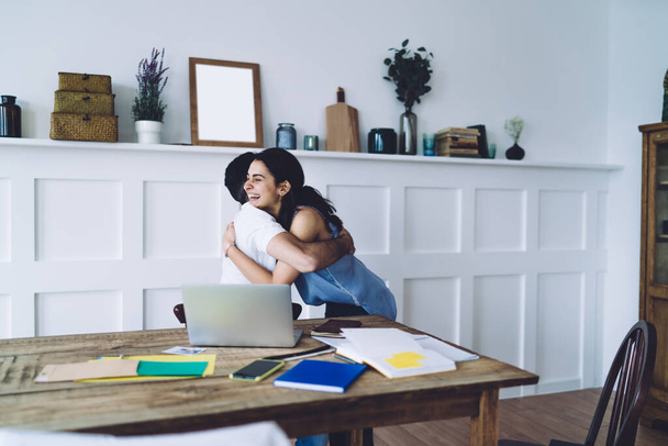 Side view of man in white T-shirt and young happy dark haired woman with toothy smile hugging and congratulating each other in modern cozy workplace - Fotoğraf, Görsel
