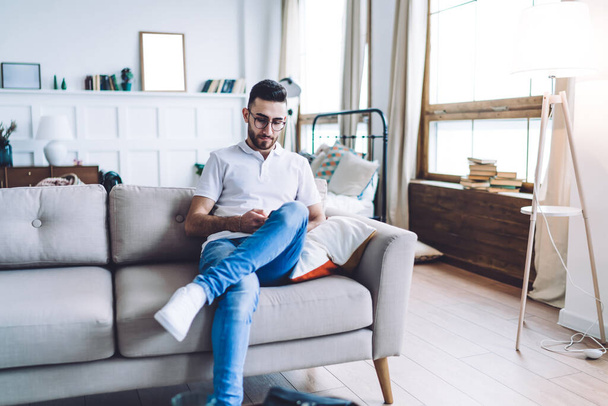 Young focused guy in jeans and white polo shirt sitting on grey comfortable sofa with legs crossed and messaging on smartphone on blurred background with living room - Fotografie, Obrázek