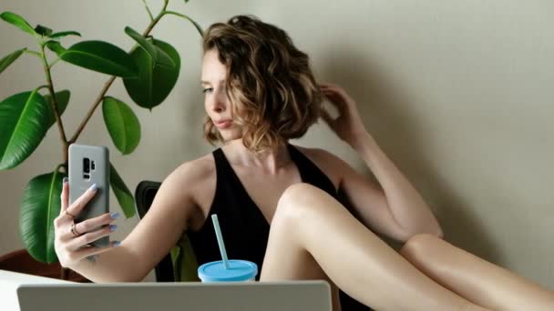 Attractive young woman smiles, poses, takes selfies and streams video to social media from her smartphone. Closeup portrait of happy woman with coktail in swimwear making photo in home office - Footage, Video