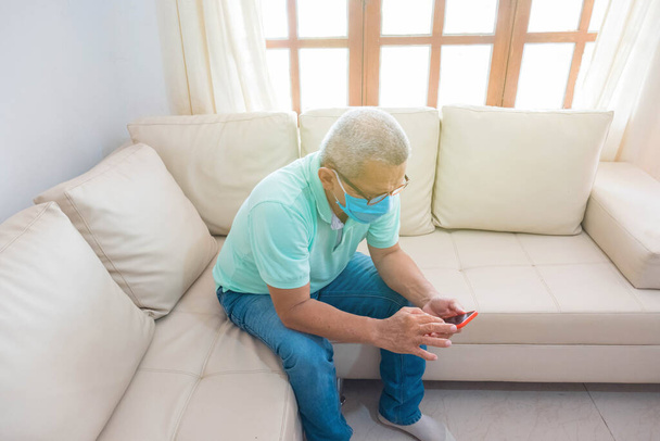 Ένας ηλικιωμένος με ιατρική μάσκα στέλνει μήνυμα στο κινητό του στο σπίτι. - Φωτογραφία, εικόνα