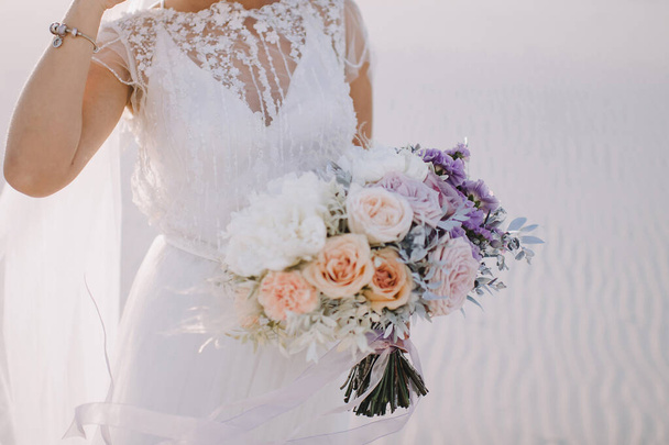 Nahaufnahme einer Bräute Strauß von Rosen, Nelken in weiß, rosa, violett, Lavendel, Pfirsichtönen. Die Braut in einem weißen Spitzenkleid hält einen Strauß in den Händen. Weißer Sand - Foto, Bild