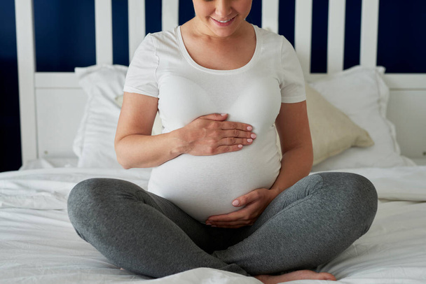 Έγκυος γυναίκα κάθεται στο κρεβάτι και κρατώντας τα χέρια στο στομάχι - Φωτογραφία, εικόνα