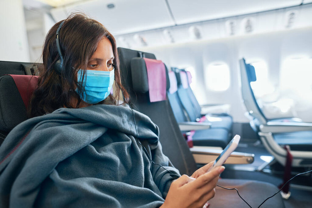Woman in virus protection face mask using smartphone sitting in empty airplane - Foto, imagen
