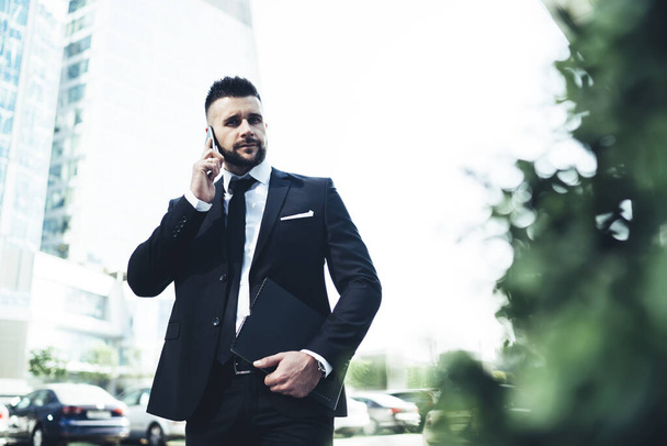 Handsome young man in black suit with notebook using cellphone for discussion with coworker freelance project walking in business district - Foto, Bild