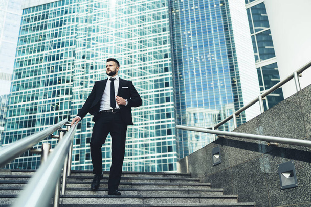 From below of successful businessman in suit walking down stairs in hurry looking away in front of giant modern business center  - Fotoğraf, Görsel