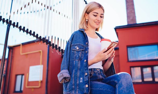 Cheerful content female in jacket and white t shirt with cup of coffee surfing on mobile phone sitting on wooden podium on roof - Фото, изображение