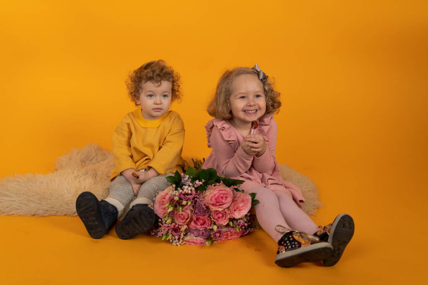 Симпатичные мальчик и девочка сидят на пушистом ковре с букет цветов, желтый фон, концепция праздника, радость и счастье в доме
 - Фото, изображение