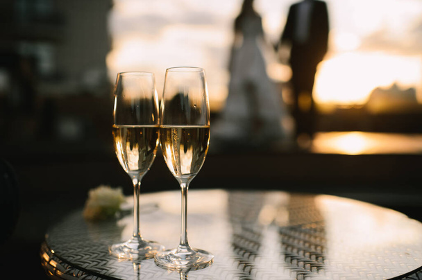 Two glasses of champagne stand on a table stand on a table. A loving couple of newlyweds is standing in the background.  - Photo, Image