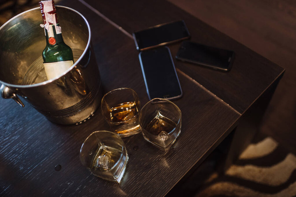 On a dark brown wooden table are three glasses of whiskey, a smartphone, an ice bucket and a bottle of whiskey. - Photo, image