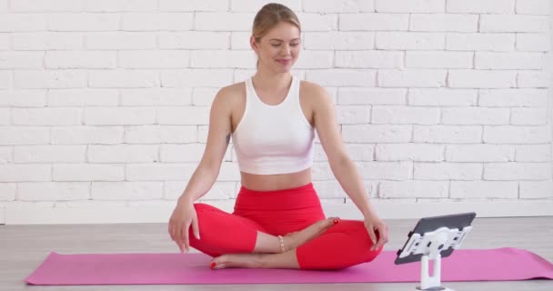 Schöne sportliche Frau sitzt mit überkreuzten Beinen auf einer Matte und schaut Yoga-Online-Unterricht - Filmmaterial, Video