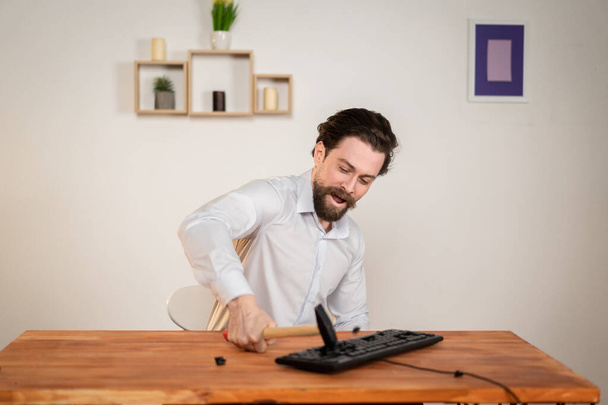 Agressieve kantoormedewerker, boze man met een baard vernietigen zijn computer, met hamer in handen vernietigen toetsenbord. - Foto, afbeelding