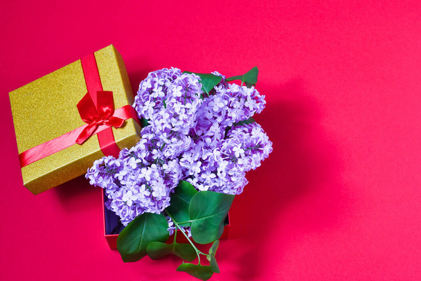 Beautiful gift box with ribbon bow and a bouquet of lilacs on a red background. Place for text. Holiday concept - Photo, Image