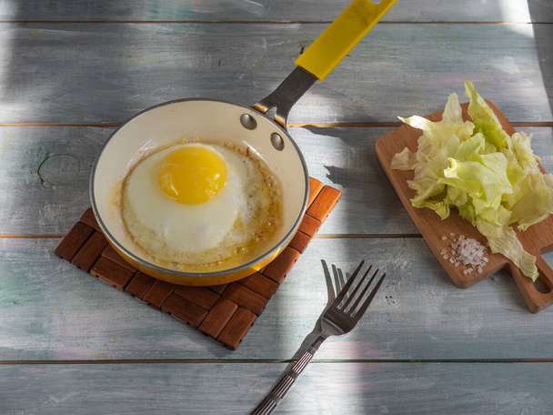 簡単な朝食、フライパンの卵1個、木製のまな板の上の氷山サラダ - 写真・画像