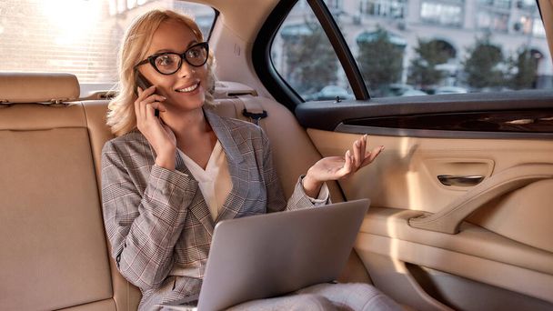 Happy to hear you. Happy and beautiful business woman in full suit working on laptop and talking on the phone while sitting in the car - Φωτογραφία, εικόνα