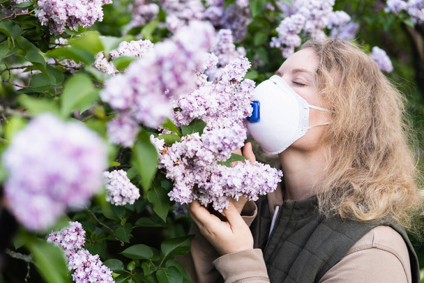 Jonge blonde vrouw ruikt lila boom bloesem door FFP2 respirator. Conceptueel beeld van corona virus quarantaine en allergie. - Foto, afbeelding