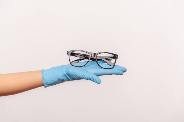 Profile side view closeup of human hand in blue surgical gloves holding and giving black eyeglasses frame. indoor, studio shot, isolated on gray background. - Foto, imagen