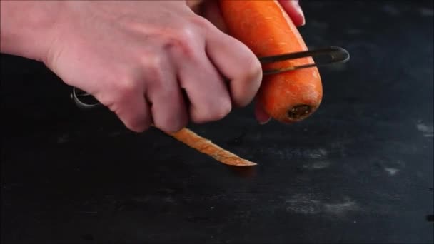 τα χέρια νεαρών γυναικών καθαρίζουν φρέσκα ολόκληρα καρότα με ένα μαχαίρι καθαρισμού, σε ένα σκούρο τραπέζι - Πλάνα, βίντεο