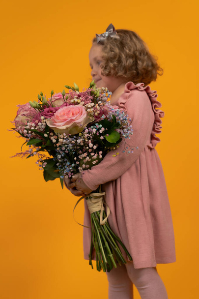 Cute little girl in a pink dress, curly hair with a bow holding a large bouquet of pink flowers in her hands, full-length profile, vertical postcard - Fotografie, Obrázek
