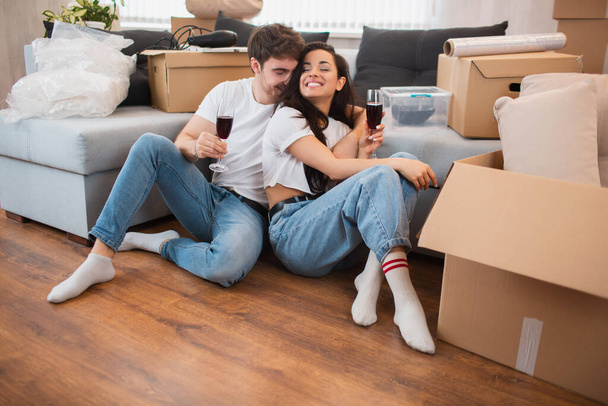 Joven pareja feliz bebiendo vino, celebrando mudarse a un nuevo hogar y sentarse entre cajas
 - Foto, imagen