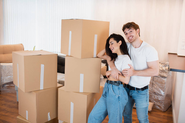 Ein junges Ehepaar steht im Wohnzimmer des Hauses neben ausgepackten Kartons. Sie freuen sich über ein neues Zuhause. Umzug, Hauskauf, Wohnungskonzept. - Foto, Bild