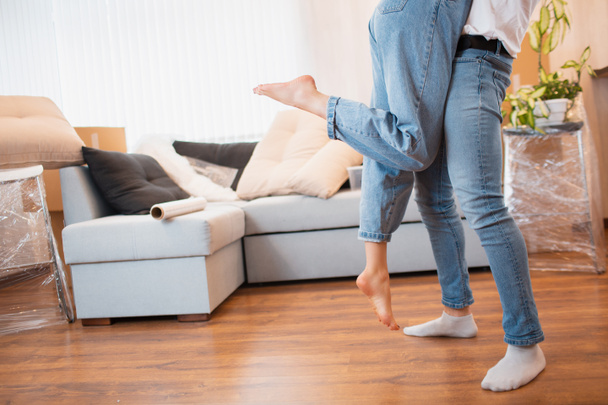 legs close up view. Young couple first time home owners celebrate moving day concept, man husband lifting holding wife standing near boxes in new own house apartment, relocation and family mortgage. - Φωτογραφία, εικόνα