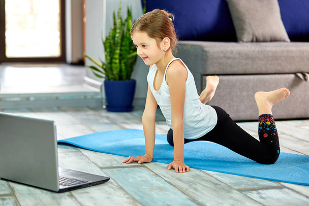 Little girl practicing yoga, stretching, fitness by video on notebook. Distant online education training, aerobic at home. Healthy lifestyle, coronavirus, stay home. Kids sport home quarantine. - Photo, Image