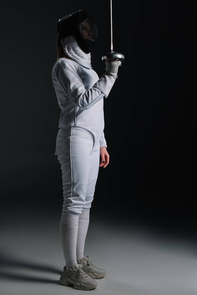 Side view of fencer in fencing suit and mask holding rapier on grey surface on black background - Photo, Image