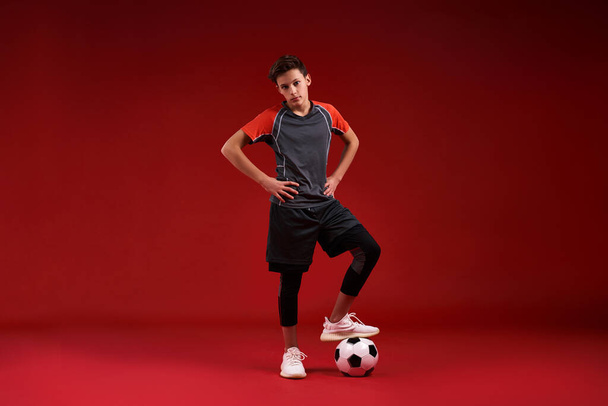 Kick it. A teenage boy is engaged in sport, he is looking at camera while exercising with football. Isolated on red background. Fitness, training, active lifestyle concept. Horizontal shot. - Fotó, kép