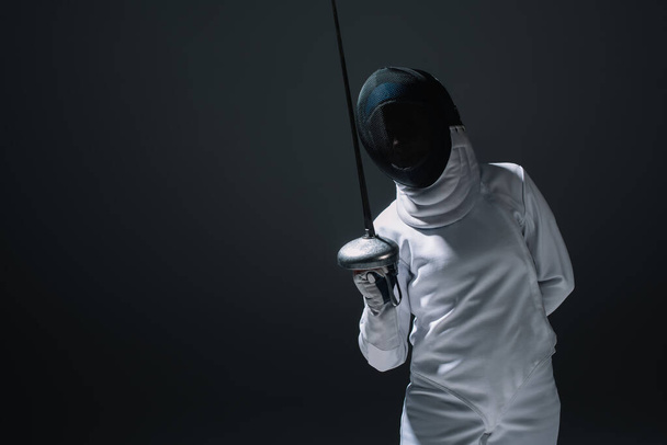 Swordswoman in fencing suit and mask holding rapier isolated on black  - Photo, Image