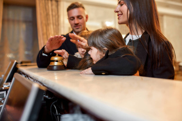 We are here. Happy family checking in hotel at reception desk. A son and a daughter are ringing a service bell together with parents while standing at the hotel reception. - Fotoğraf, Görsel