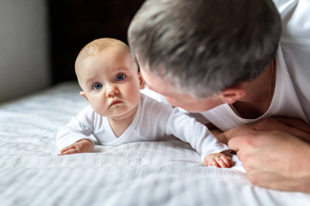 Pretty baby with dad on a white sheet on the bed. Cute infant child with father. Can be used in journal. Newborn baby and dad, a newborn baby in her husband's arms. Father with son or daugher - Фото, зображення