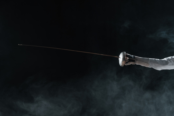 Cropped view of fencer in glove holding rapier on black background with smoke  - Foto, Bild