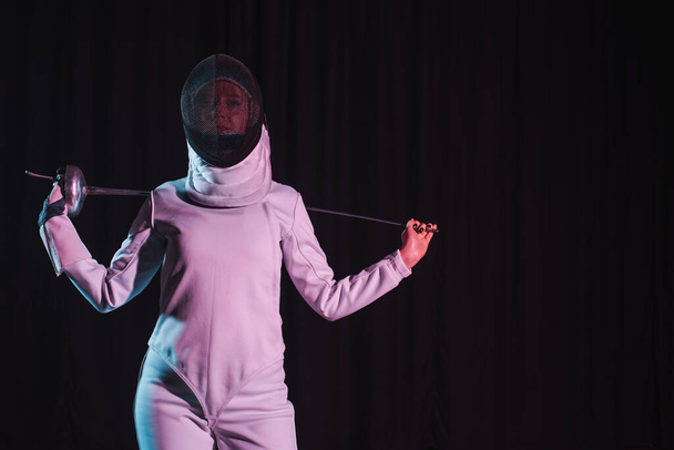 Swordswoman in fencing mask holding rapier isolated on black with copy space - Valokuva, kuva
