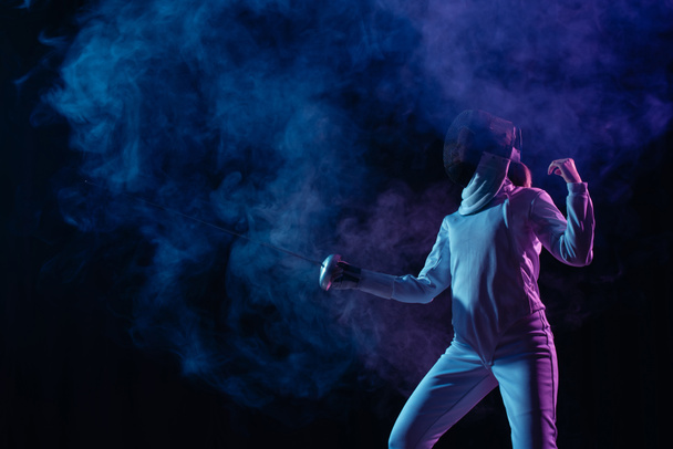 Fencer exercising with rapier on black background with smoke  - Foto, Imagen