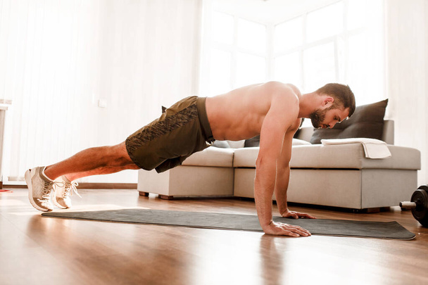 Young man go in for sport at home. Full size picture of t-shirtless guy sportsman stand in plank position alone in room. Beginner try to do his best and exercise. Hardworking real person - Photo, Image