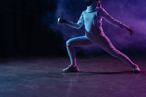 Cropped view of swordswoman fencing on black background with smoke and lighting  - Foto, immagini