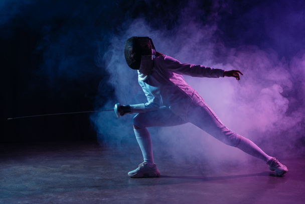 Swordswoman fencing with rapier on black background with colorful smoke  - Photo, Image