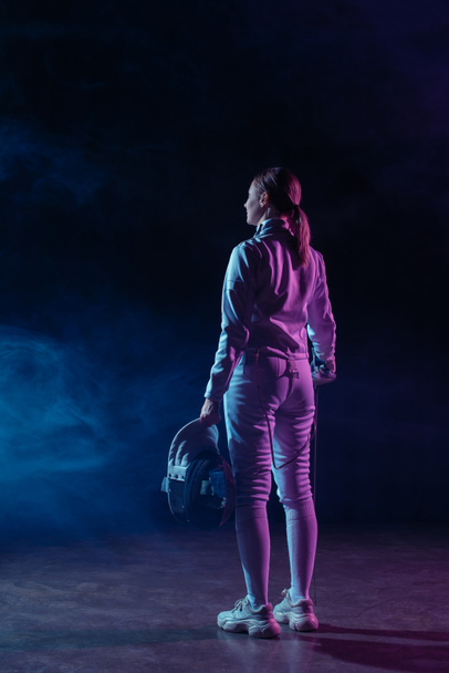 Back view of fencer holding fencing mask and rapier on black background with smoke  - Foto, Bild