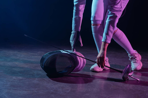 Cropped view of swordswoman putting fencing mask and rapier on floor on black background - Photo, Image